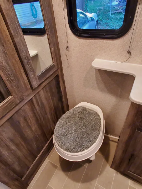 RV rental bathroom featuring a toilet with a gray shag cover, wood paneling, white shelf, and outside views from windows.. Forest River Salem 2020