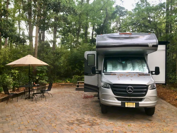 Silver Mercedes-Benz RV rental at a wooded campsite with an outdoor table, chairs, and umbrella setup.. Winnebago Mercedes Sprinter 2020