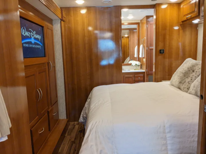 This RV rental bedroom offers a comfortable bed, a TV showing Disney, and an open doorway to the connected bathroom with a vanity.. Coachmen Mirada 2019