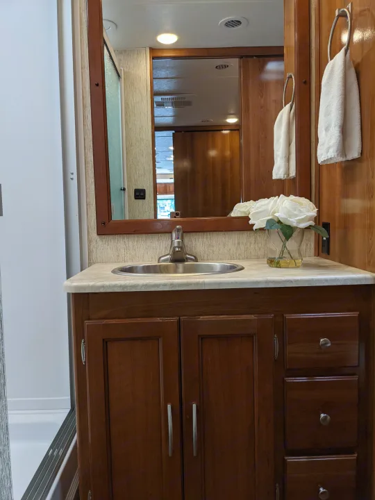 RV rental bathroom with a compact wooden vanity, sink, mirror, white flowers, and towel. Reflects interior space.. Coachmen Mirada 2019