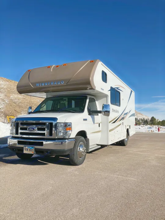 White Winnebago Minnie Winnie RV rental parked on asphalt, with snowy hills and a clear blue sky.. Winnebago Minnie Winnie 2016