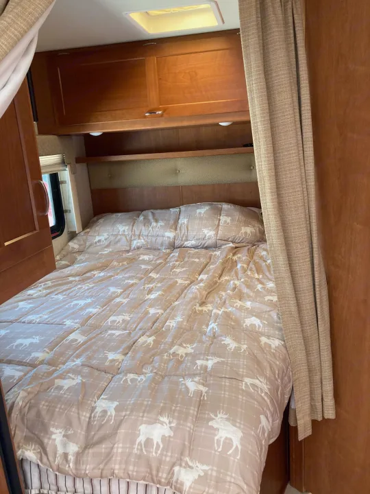 RV rental's cozy bedroom: a bed with light brown moose-patterned comforter, wood cabinets, and window.. Winnebago Minnie Winnie 2016
