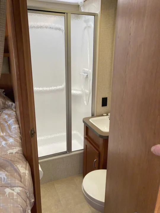 This RV rental interior showcases a compact bathroom with a shower, sink, and toilet, adjacent to a bed with deer-patterned bedding.. Winnebago Minnie Winnie 2016