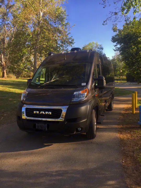 A dark grey Ram ProMaster RV rental is parked on an asphalt path in a sunny park, surrounded by lush trees and grass.. Thor Sequence 2021