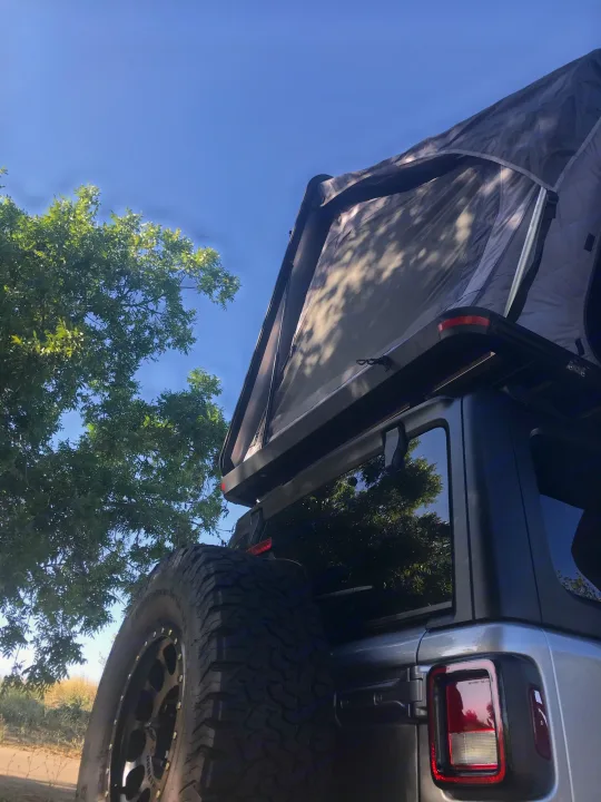 A grey Jeep with an open rooftop tent for **rv rental** adventures. Perfect for camping under the blue sky near green trees.. Other Other 2019