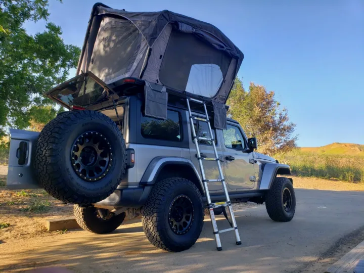 Silver Jeep Wrangler with a deployed rooftop tent and ladder setup, ready for adventure camping. A unique RV rental option.. Other Other 2019