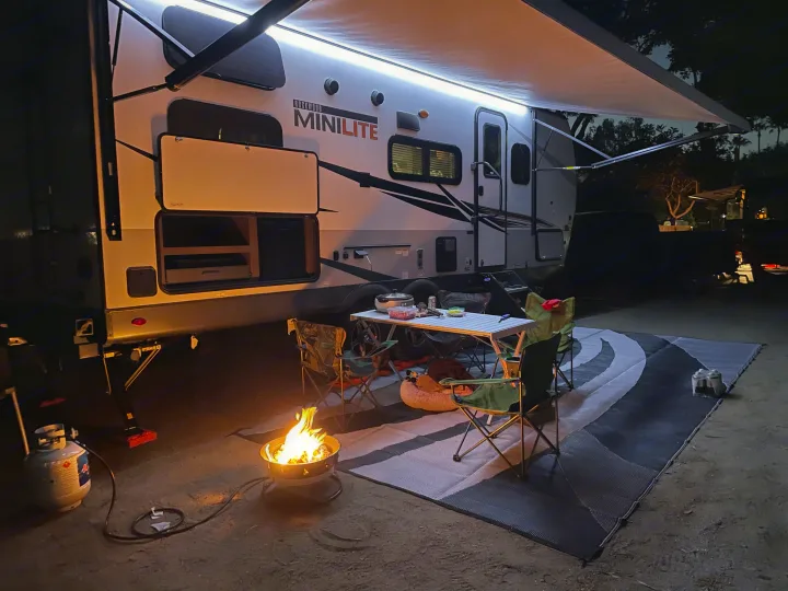 Experience comfortable camping with this RV rental. Evening scene with lit awning, table, chairs, fire pit.. Forest River Mini-Lite 2021