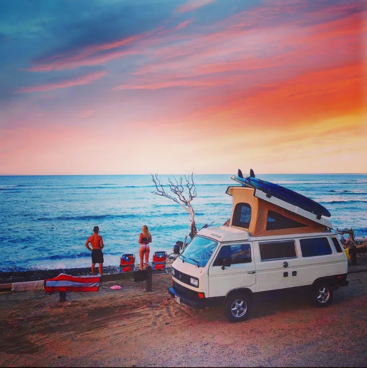 RV rental dream: White camper van with surfboards parked beachfront at stunning sunset. Couple enjoys ocean view.. VW Westfalia 1986