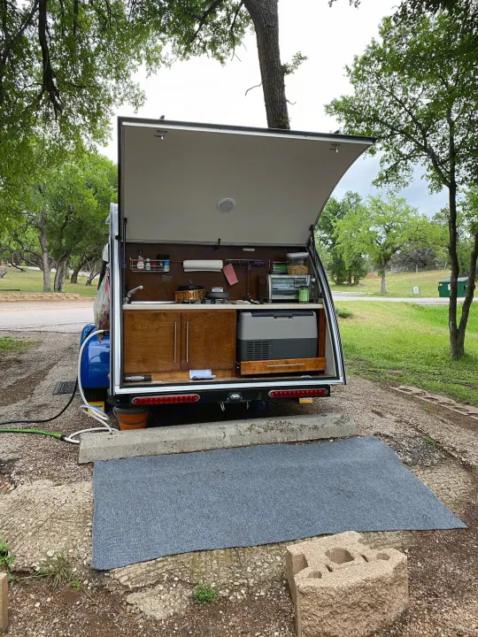 This RV rental shows an open rear kitchen setup with a sink, fridge, and toaster oven at a wooded campsite, ideal for outdoor cooking.. Little Guy NuCamp 2015