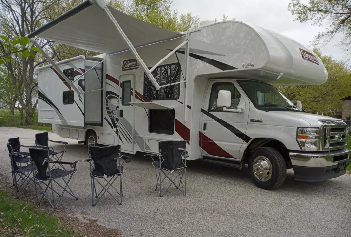 Ready for adventure: a white Coleman rv rental motorhome with an extended awning, outdoor TV, and camping chairs set up.. Coleman Thor 2021