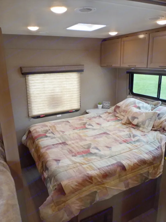 Cozy RV rental bedroom featuring a patterned bed, compact layout, overhead cabinets, and inviting windows.. Coleman Thor 2021