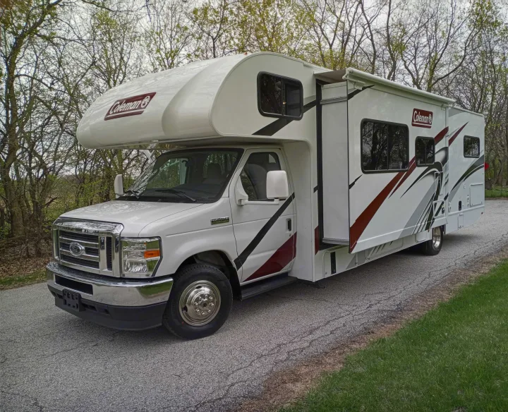 White Coleman Class C RV rental with graphic stripes, awning extended & slide-out open, parked on a road.. Coleman Thor 2021