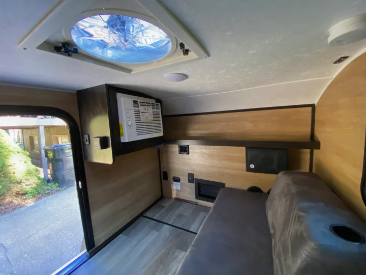 RV rental interior view: ceiling fan, wall-mounted AC, and cushioned bench seat. An open door shows the outdoors.. Bushwacker 10SS 2021