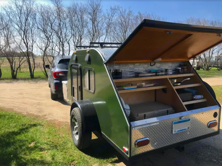 Green teardrop RV rental attached to a car, with its rear hatch open revealing a compact outdoor kitchen and storage.. American Teardrop Travel Trailer 2016