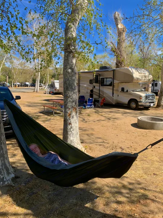 A young woman relaxes in a green hammock, surrounded by trees, with an RV rental and campsite in the sunny background.. Other Other 2010