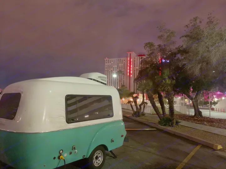Night view of a retro **rv rental** camper, white & turquoise, parked at Circus Circus Las Vegas. Bright neon sign and trees visible.. Happier Camper HC1 2020