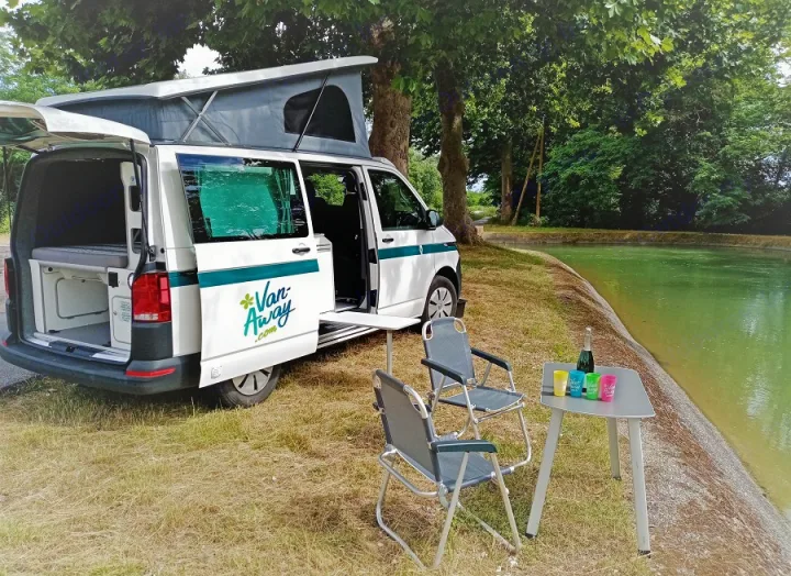 Experience freedom with this RV rental! A camper van with open pop-top, chairs, and table by a tranquil canal.. Volkswagen Transporter T6 2021