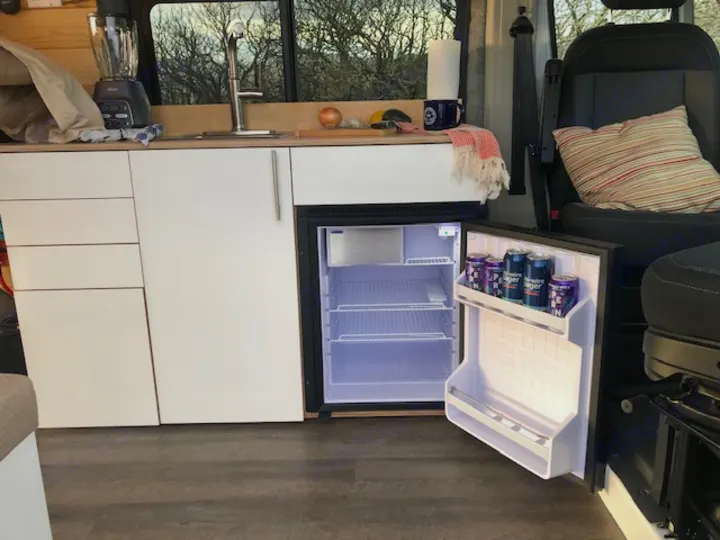 Inside your RV rental: a compact kitchen featuring a bright, open fridge, sink, and clean white cabinets.. Ram Promaster 2021