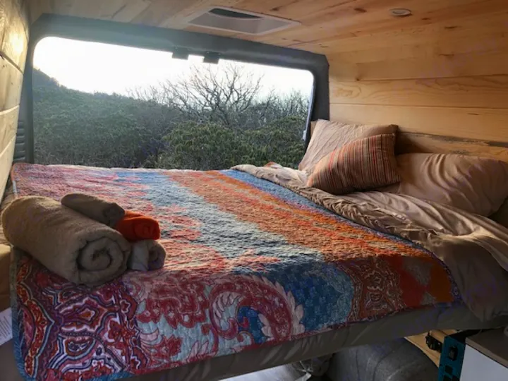 Comfortable rv rental bed with colorful quilt, rolled towels, and a beautiful natural view from the large window.. Ram Promaster 2021
