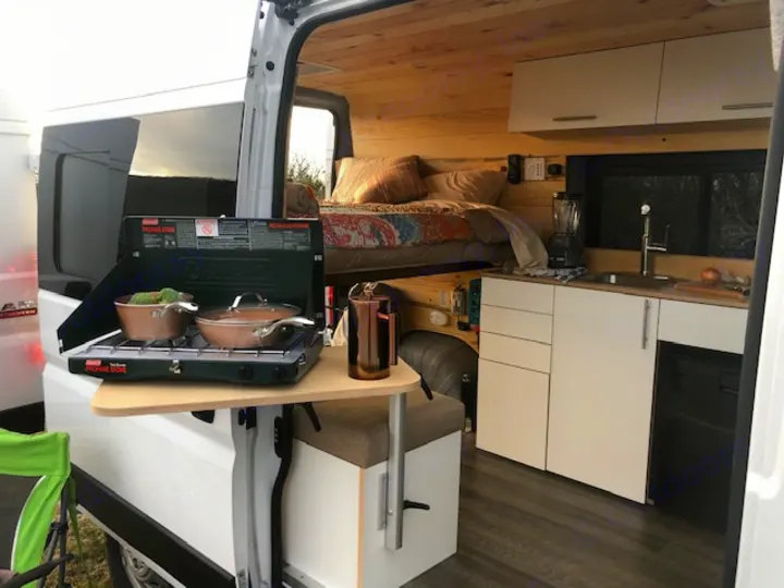 This rv rental features a cozy interior with a built-in kitchen, elevated bed, and outdoor propane stove setup for versatile camping.. Ram Promaster 2021