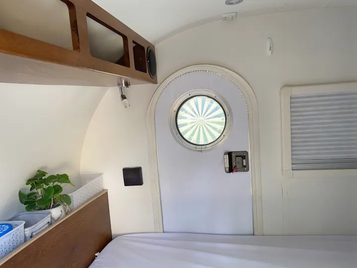 Bright rv rental interior. Features a unique porthole door, bed, wooden cabinets, and a potted plant on a built-in shelf.. Little Guy NuCamp 2015