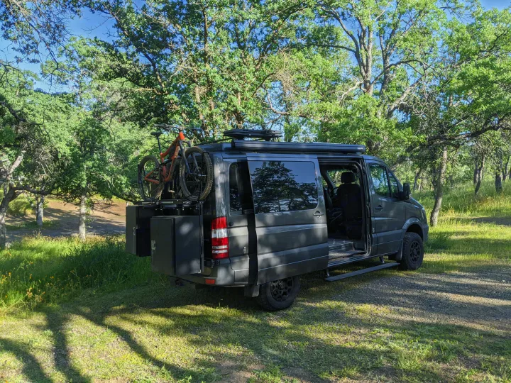 Explore the outdoors with this gray camper van RV rental, equipped with a mountain bike, parked under trees.. Mercedes-Benz Sprinter 2016
