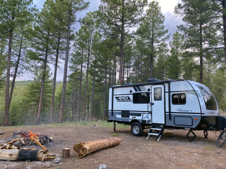 An RV rental is set up in a lush pine forest, featuring a warm campfire. Ideal for a tranquil outdoor camping trip.. Coachmen Apex 2021