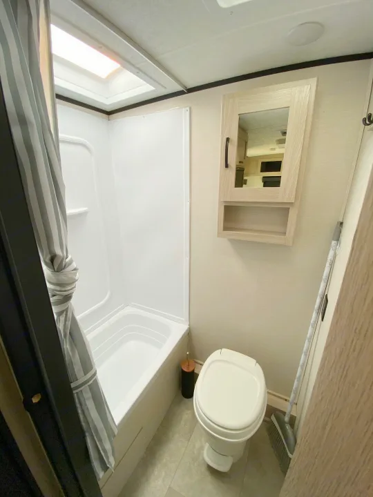 Inside an RV rental: a compact bathroom with shower/tub, toilet, and mirrored cabinet, illuminated by a skylight.. Coachmen Apex 2021