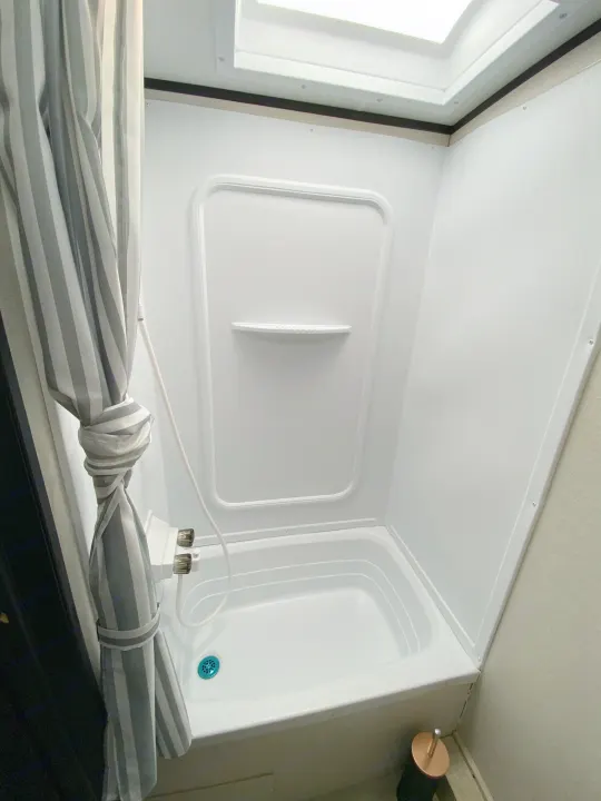 Inside an RV rental, this bright white shower unit features a tied-back striped curtain, built-in shelf, and blue drain.. Coachmen Apex 2021