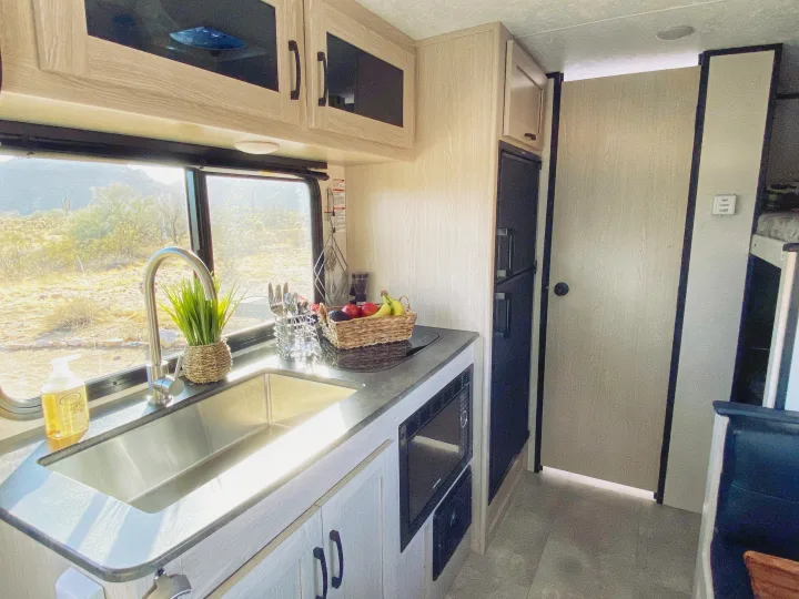 Modern **RV rental** kitchen featuring a stainless steel sink, microwave, fridge, and desert landscape through window.. Coachmen Apex 2021