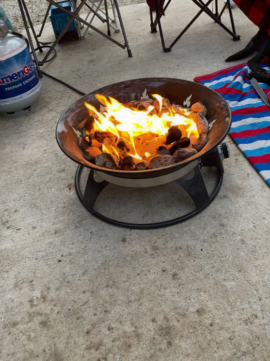 Enjoy your RV rental trip! A portable propane fire pit with flickering flames and lava rocks sits on concrete, next to camping chairs.. Forest River Wildwood 2021