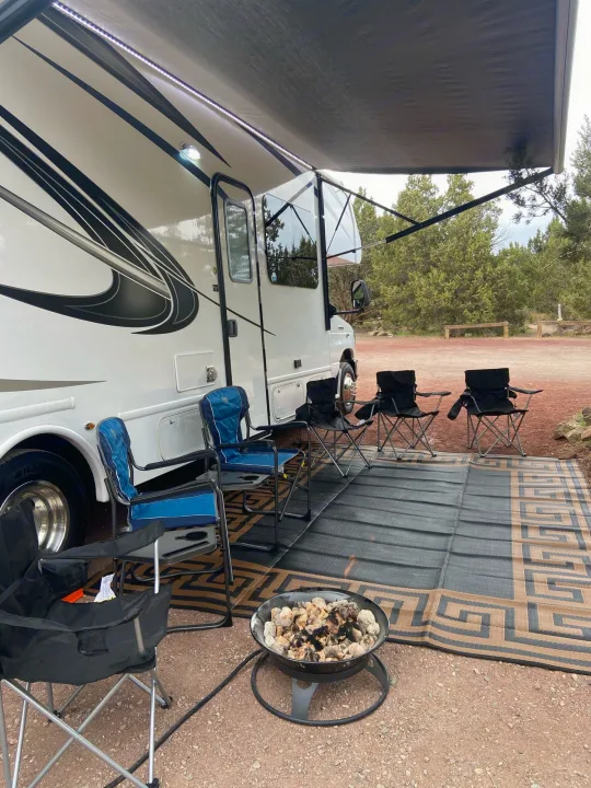 Relaxing RV rental setup: an awning-covered campsite with chairs, a mat, and a fire pit, surrounded by nature.. Nexus Triumph 2021