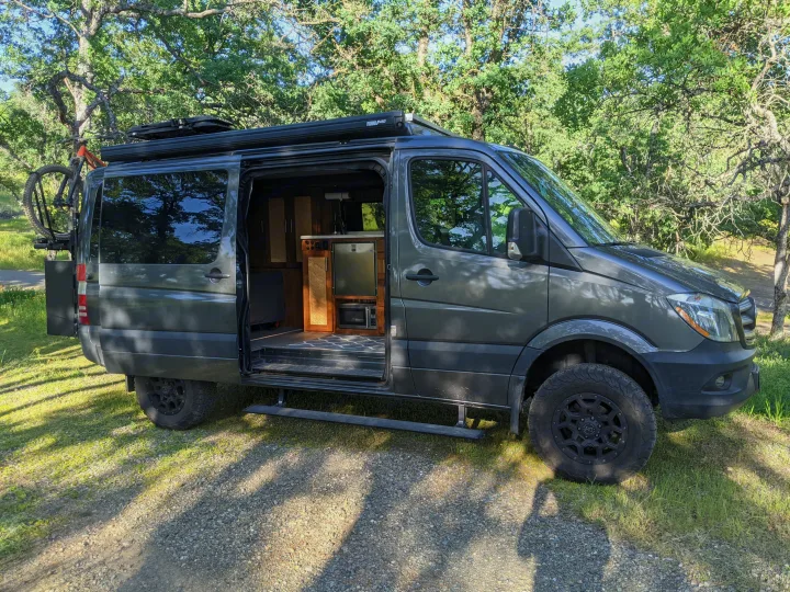 Explore in this rugged dark gray Sprinter RV rental camper van, featuring an open door revealing its interior, bike rack, & off-road tires.. Mercedes-Benz Sprinter 2016