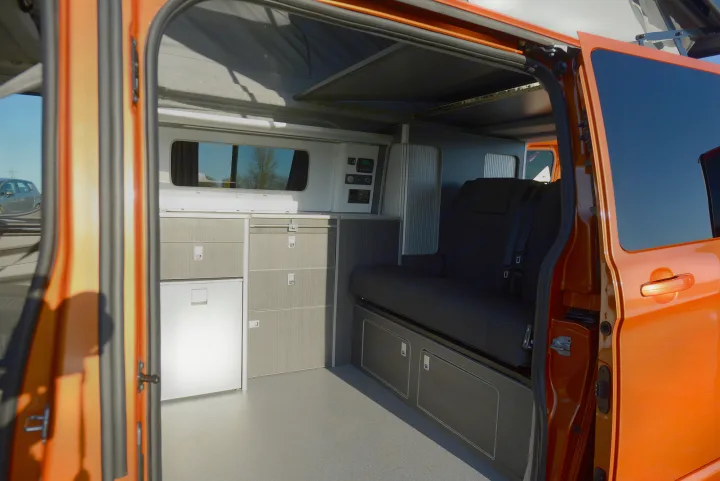 Orange camper van interior for RV rental. Shows kitchen unit with fridge, control panel, and rear bench seating.. Ford Custom 2018