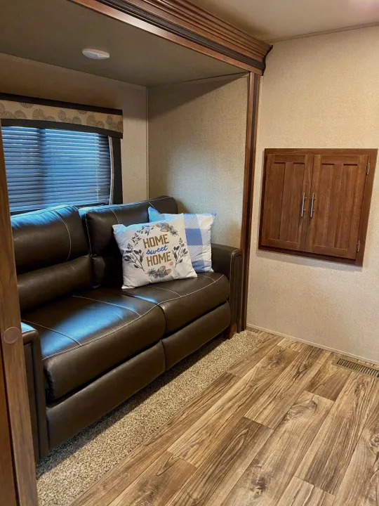 Cozy RV rental interior: brown sofa, "HOME sweet HOME" pillow, window, wood-look floor, and wood wall cabinet.. Keystone Volante 3801MD 2018