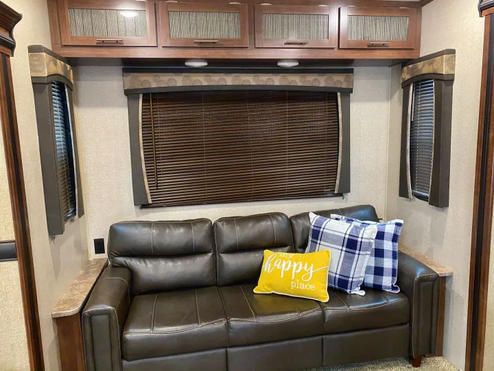 Comfortable RV rental living area featuring a dark leather sofa with throw pillows, wooden cabinets, and window blinds.. Keystone Volante 3801MD 2018