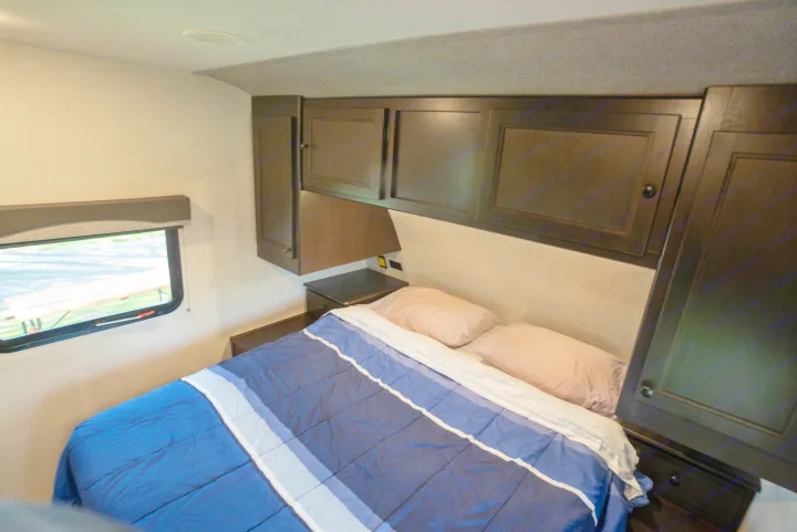 Cozy RV rental bedroom with a blue striped bed, dark wood overhead cabinets, and a window, ideal for travel.. Starcraft Super Lite 2021