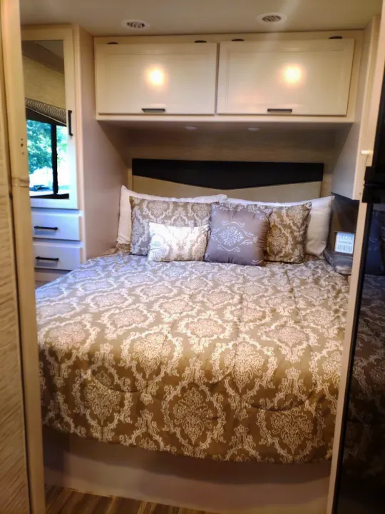 Cozy RV rental bedroom featuring patterned bedding, overhead cabinets, and a window, ideal for comfortable travel accommodations.. Thor Four Winds 2021
