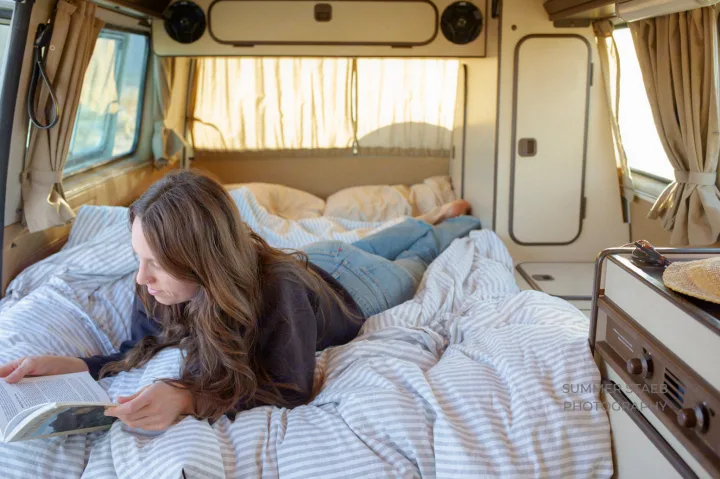 Woman relaxes in an RV rental, reading a book on a striped bed inside a cozy camper van interior.. Volkswagen Westfalia 1986