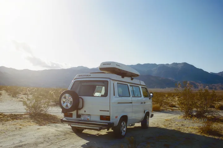 White Westfalia camper van with roof cargo box, parked in a sunny desert landscape with mountains; ideal RV rental.. Volkswagen Westfalia 1986