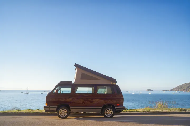 Experience coastal beauty! Brown pop-top camper van by a sunny bay with sailboats. Explore with an RV rental.. Volkswagen Westfalia 1991