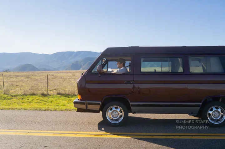 Scenic road trip: an RV rental (maroon camper van) driven by a man, passing fields & mountains on a sunny day.. Volkswagen Westfalia 1991