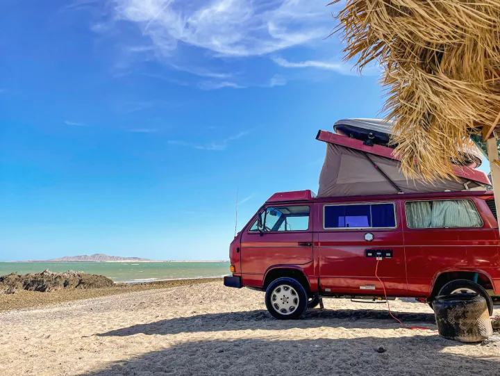 Explore by rv rental: Red camper van parked on a sandy beach by the ocean under a clear blue sky, pop-top deployed.. Volkswagen Westfalia 1987