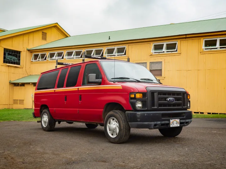 A vibrant red Ford E-Series camper van, an RV rental with Dakine racks, parked by a sunny yellow Hawaiian building.. Ford E150 2011