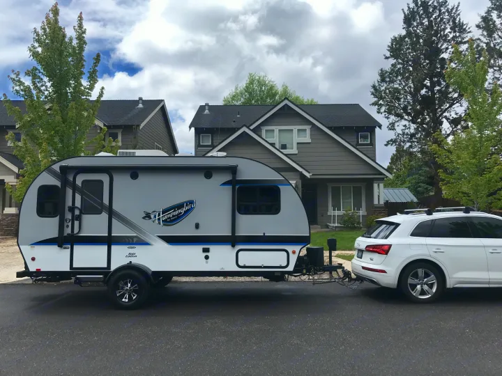 A white SUV hitches an RV travel trailer, perfect for your next RV rental road trip, parked on a residential street.. Jayco Baja 2018