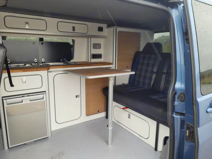 RV rental camper van interior with kitchenette, fridge, adjustable table, and plaid bench seating next to an open door.. Volkswagen T5 2018