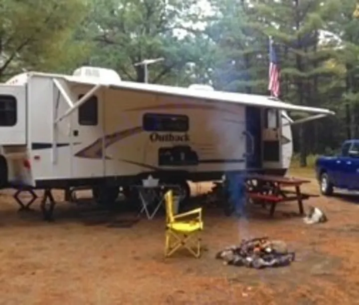 This RV rental is set up at a forest campsite. An extended awning covers a picnic table & chairs near a glowing campfire for outdoor fun.. Keystone RV Outback 2011