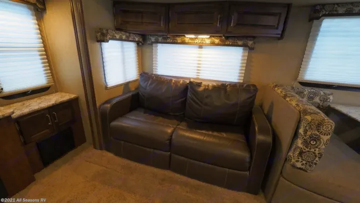 Inside an RV rental, this cozy living space has a brown leather sofa, wood cabinets, and bright windows with pleated blinds.. Keystone Outback 2016