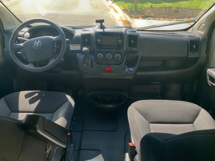 View of an RV rental van's interior, showing the detailed dashboard, steering wheel, and front seats in sunlight.. Winnebago Winnebago 2021