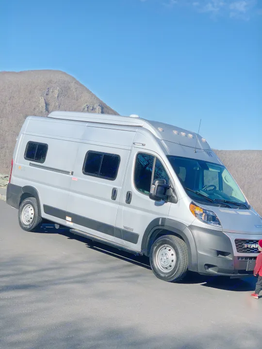 Silver Winnebago Solis camper van, an RV rental, parked on asphalt with mountains under a clear blue sky. A child is nearby.. Winnebago Winnebago 2021